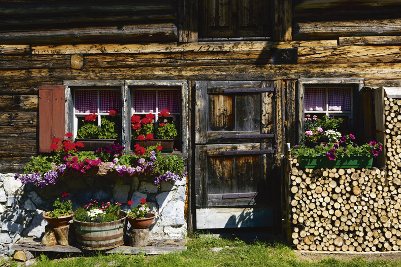 Man sieht den Eingang eines alten Holzbauernhofes, geschmückt  mit Blumen und einer Scheitertriste.