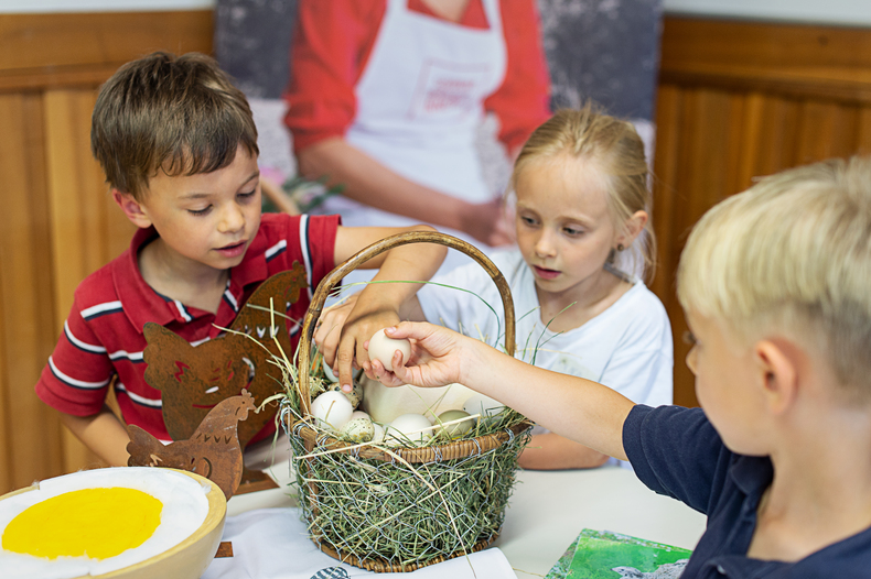 Drei Kinder sitzen an einem Tisch, vor einem mit Heu und Eier gefüllten Korb und nehmen ein Ei heraus.