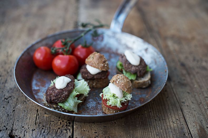 In einer Eisenpfanne sind 4 mit Fleisch, Salat und Käse gefüllten Miniburger mit 4 Tomaten angerichtet.