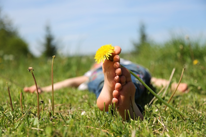 Eine Person liegt im grünen Gras, die Füße übersschränkt, in der Zehe steckt eine Löwenzahnblüte. Eine Person liegt im grünen Gras, die Füße übersschränkt, in der Zehe steckt eine Löwenzahnblüte.